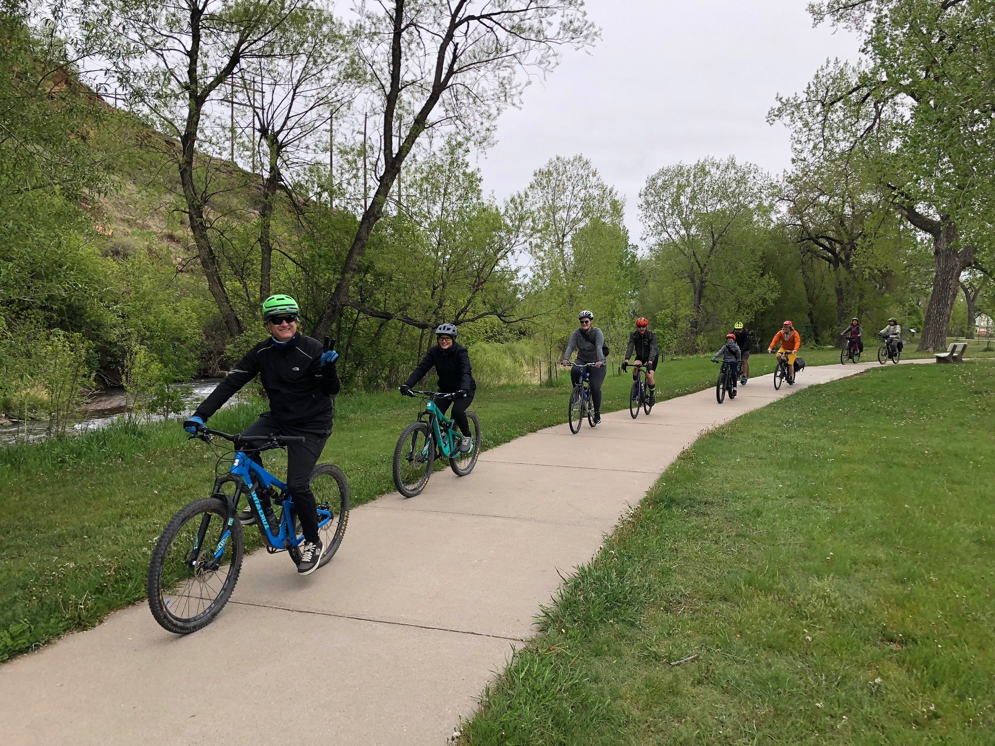 A group of cyclists in helmets riding along a paved path beside a creek